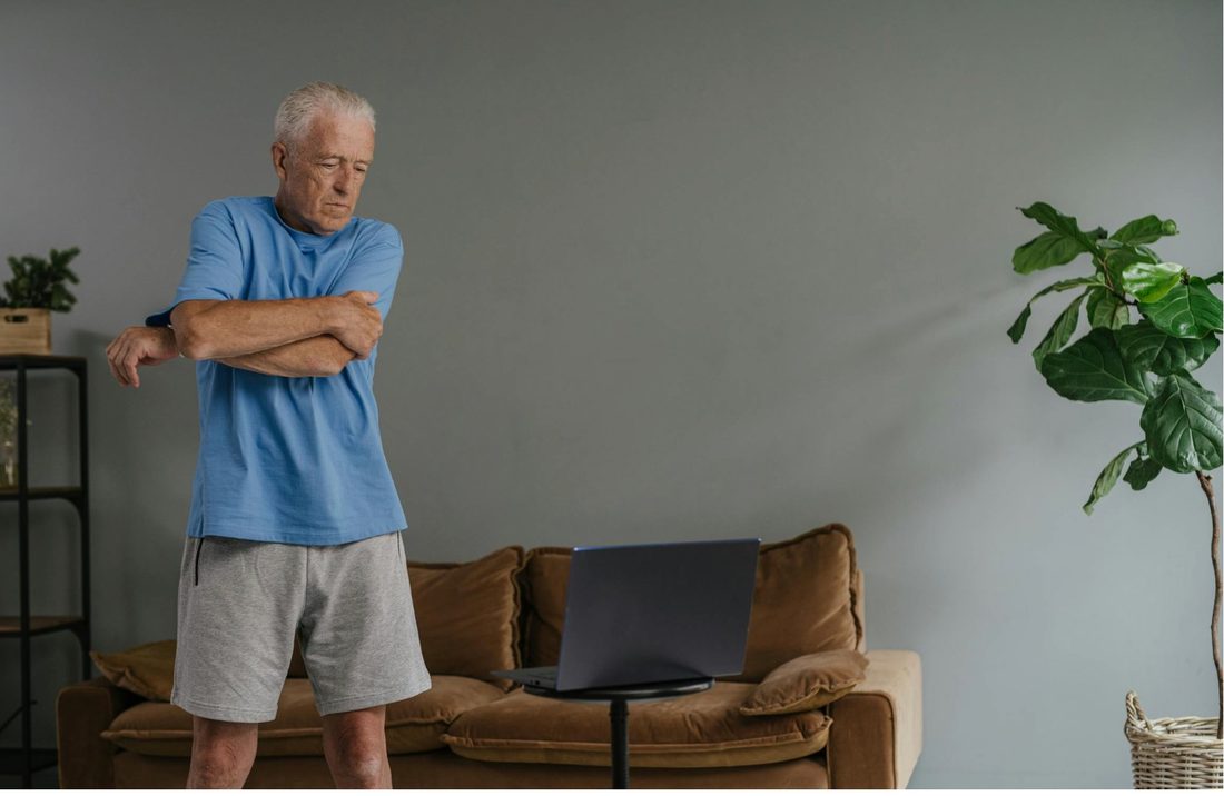 An older man stretching his arm during a guided exercise session at home in front of a laptop