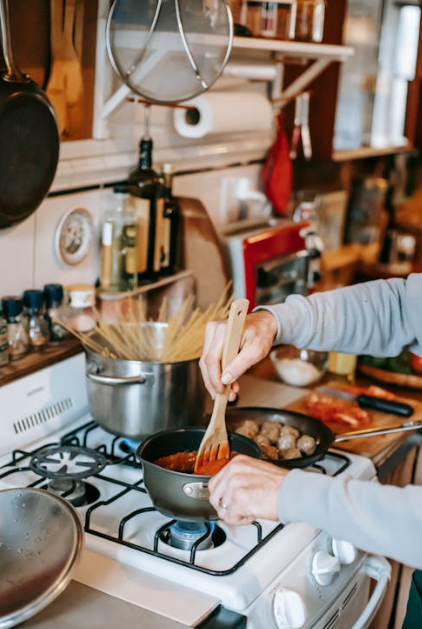 A person cooking pasta on a home gas stove, stirring a small pan of sauce next to a pot of boiling water