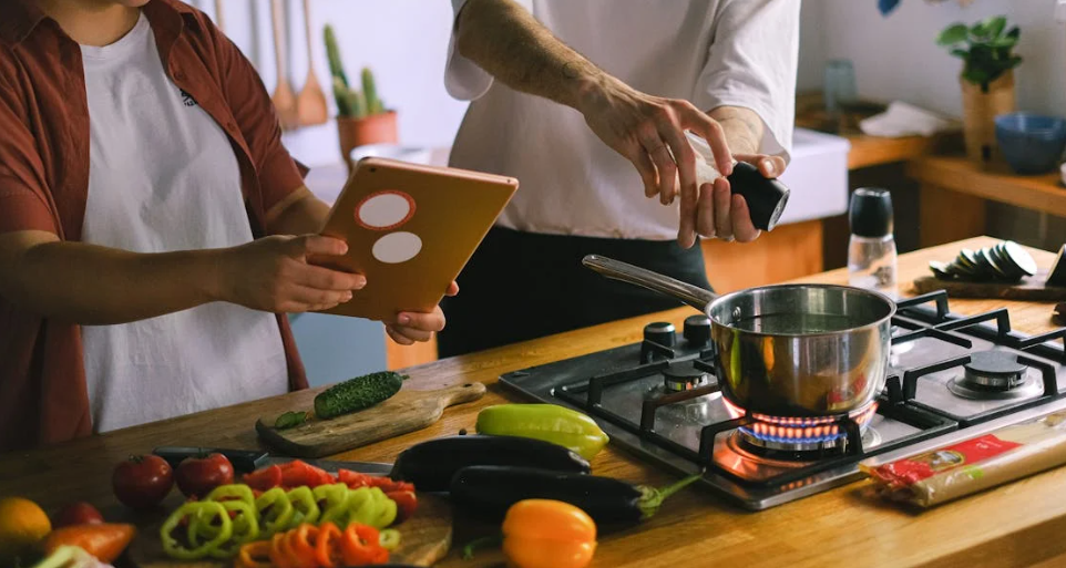Two people cooking together at a sunlit kitchen counter, chopping vegetables and stirring a pot on the stove