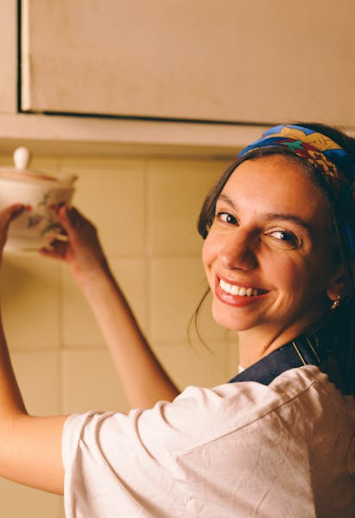 A person smiling in a home kitchen, holding a small ceramic pot, wearing a colorful patterned headscarf